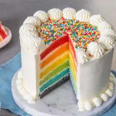 Torta arcobaleno con strati colorati, glassa bianca e zuccherini. Una fetta mostra i colori vivaci.