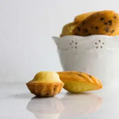 Due madeleine dolci, una a forma di conchiglia e una più tondeggiante, su un piano di marmo. Sullo sfondo, una ciotola bianca piena di altre madeleine.