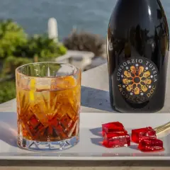 Un bicchiere di spritz con spumante, una bottiglia di Prosecco e cubetti di gelatina rossa su un vassoio bianco, con vista sul mare.