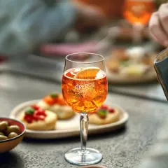 Un bicchiere di Aperol Spritz, ghiaccio e una fetta d'arancia, con olive e stuzzichini sullo sfondo.