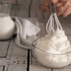 Una mano monta la panna in una ciotola di vetro. Sullo sfondo, una brocca di latte e un tovagliolo bianco.