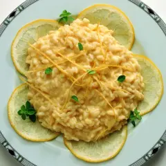 Un cremoso risotto al limone, guarnito con scorzette e foglioline di menta, circondato da fette di limone fresco.