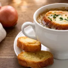 Una tazza di zuppa di cipolle fumante, con crostini dorati e formaggio fuso, accanto a una cipolla cruda e un tovagliolo di lino.