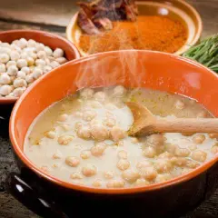 Una fumante zuppa di ceci in una ciotola arancione, con ceci secchi, spezie e rosmarino sullo sfondo.