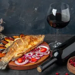 Un delizioso pesce fritto su un piatto con melanzane, cipolle rosse e pomodorini, accompagnato da vino rosso.