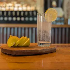 Un bicchiere di acqua frizzante con una fetta di limone, pronto per diventare la base perfetta per un rinfrescante spritz. Accanto, spicchi di limone su un tagliere di legno.