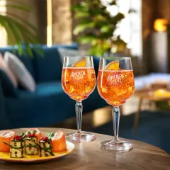 Due bicchieri di cocktail con Aperol Spritz, ghiaccio e fetta d'arancia, accompagnati da stuzzichini su un tavolino in legno.