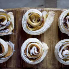 Sei rose di pasta sfoglia con mele e zucchero di canna, cosparse di zucchero a velo, su un tagliere di legno.