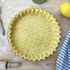 Base di pasta frolla pronta in teglia, forata e con bordi ondulati, accanto a un limone e un canovaccio a righe.