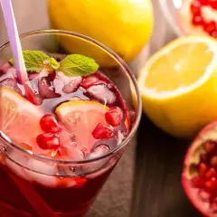 Un rinfrescante spritz al melograno con fette di limone, chicchi rossi e foglie di menta, accompagnato da limoni e melograni freschi.