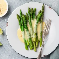 Asparagi freschi conditi con una cremosa salsa olandese, accompagnati da spicchi di lime e una forchetta d'argento.