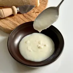 Un cucchiaio versa una densa crema di parmigiano in una ciotola. Sul tagliere, un pezzo di formaggio e un coltellino.