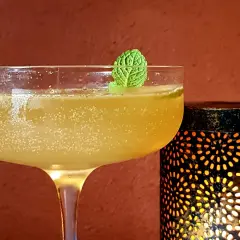 Rinfrescante cocktail con rum e menta, guarnito con una fogliolina verde, accanto a una lanterna decorativa che emana luce calda.