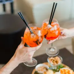 Due mani brindano con due spritz pieni di ghiaccio e cannucce nere. Sul tavolo, stuzzichini invitanti.