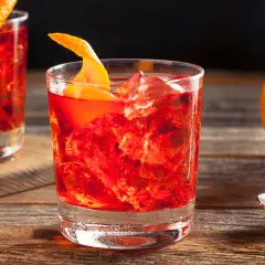 Un bicchiere di rosso vibrante, ghiaccio e scorza d'arancia. La ricetta americano è pronta per essere gustata, con un'arancia sullo sfondo.