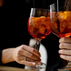 Due mani brindano con due spritz, un superalcolico rinfrescante con ghiaccio e arancia, su sfondo scuro.