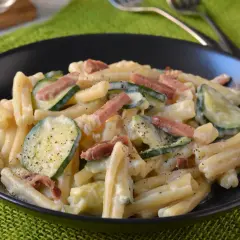 Piatto di pasta cremosa con zucchine a rondelle e cubetti di speck croccante, guarnito con pepe nero.