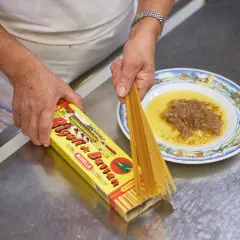 Mani che preparano i bigoli in salsa, con la scatola di pasta e un piatto di salsa di acciughe e cipolle.