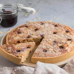 Torta con crema frangipane, mandorle a lamelle e ciliegie, spolverata di zucchero a velo. Accanto, un vasetto di confettura.