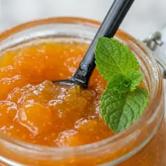 Vasetto di vetro pieno di marmellata di albicocche fatta in casa, con un cucchiaio nero e una fogliolina di menta fresca.