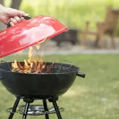Mano solleva il coperchio rosso di un barbecue a carbonella con fiamme vive. Perfetto per chi vuole sapere se posso usare legna nel barbecue a carbonella.