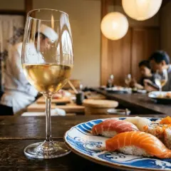 Un bicchiere di vino bianco accanto a un piatto di sushi misto, con chef e clienti sullo sfondo. Un perfetto abbinamento vino sushi per una cena raffinata.