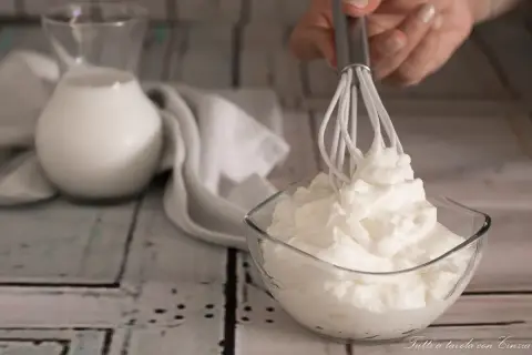 Una mano monta la panna in una ciotola di vetro. Sullo sfondo, una brocca di latte e un tovagliolo bianco.