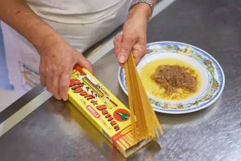 Mani che preparano i bigoli in salsa, con la scatola di pasta e un piatto di salsa di acciughe e cipolle.