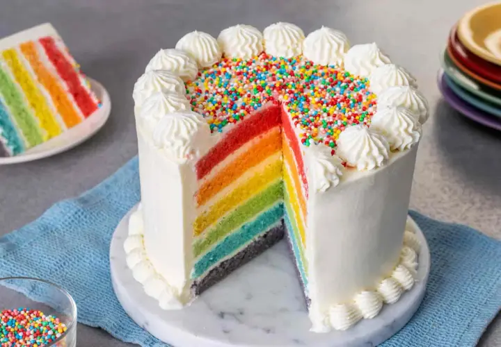 Torta arcobaleno con strati colorati, glassa bianca e zuccherini. Una fetta mostra i colori vivaci.