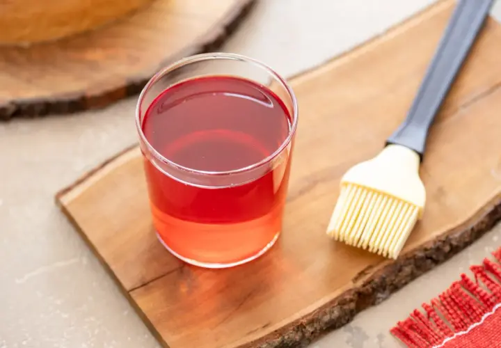 Bagna all'alchermes in un bicchiere, pronta per essere usata con un pennello da cucina su un tagliere di legno.