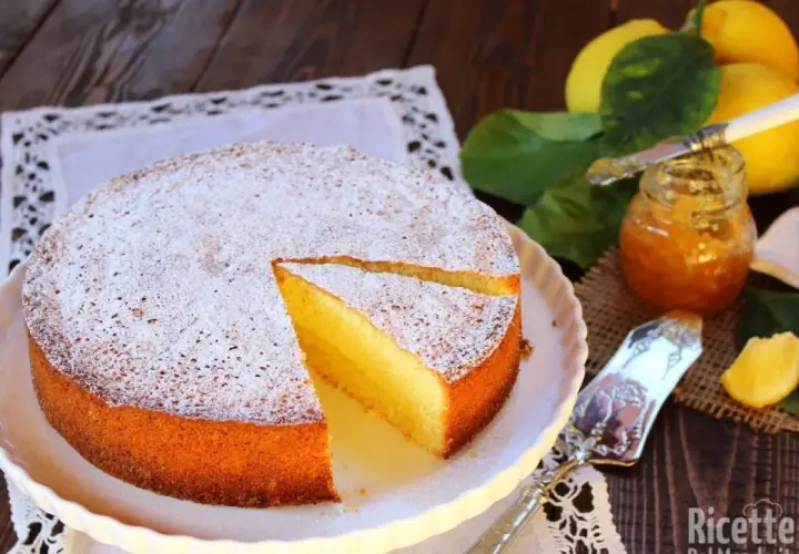 Una soffice torta al limone, spolverata di zucchero a velo, accanto a limoni freschi e marmellata.