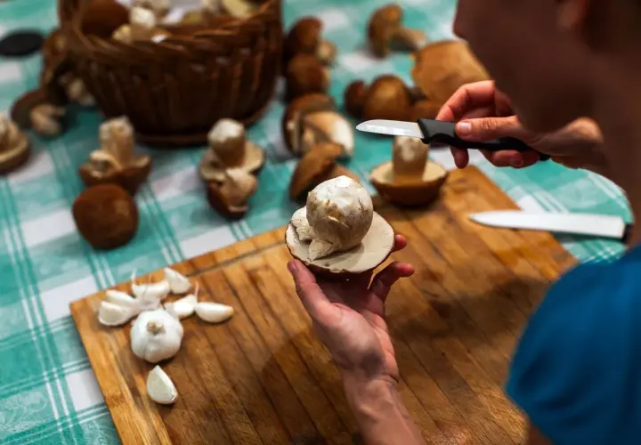 Mani che puliscono funghi freschi su un tagliere di legno, con aglio e un cesto di funghi sullo sfondo. Impara come pulire i funghi per gustarli al meglio.