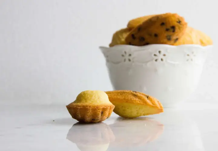 Due madeleine dolci, una a forma di conchiglia e una più tondeggiante, su un piano di marmo. Sullo sfondo, una ciotola bianca piena di altre madeleine.