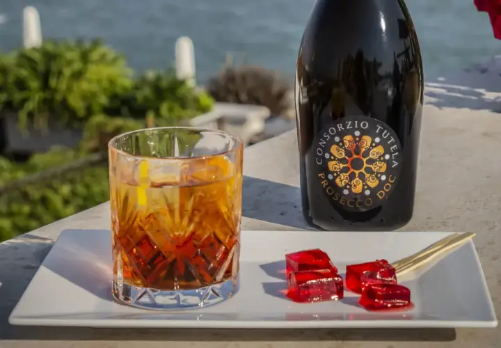 Un bicchiere di spritz con spumante, una bottiglia di Prosecco e cubetti di gelatina rossa su un vassoio bianco, con vista sul mare.