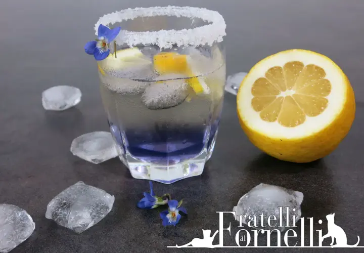 Rinfrescante liquore alla violetta spritz, guarnito con limone e fiori, circondato da cubetti di ghiaccio.