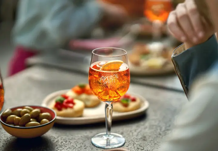 Un bicchiere di Aperol Spritz, ghiaccio e una fetta d'arancia, con olive e stuzzichini sullo sfondo.