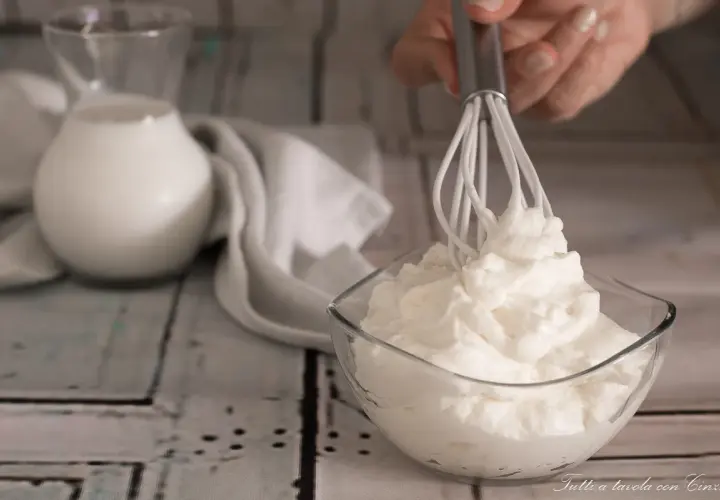Una mano monta la panna in una ciotola di vetro. Sullo sfondo, una brocca di latte e un tovagliolo bianco.