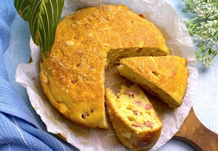 Torta salata prosciutto e formaggio dorata, con una fetta tagliata, su carta da forno.