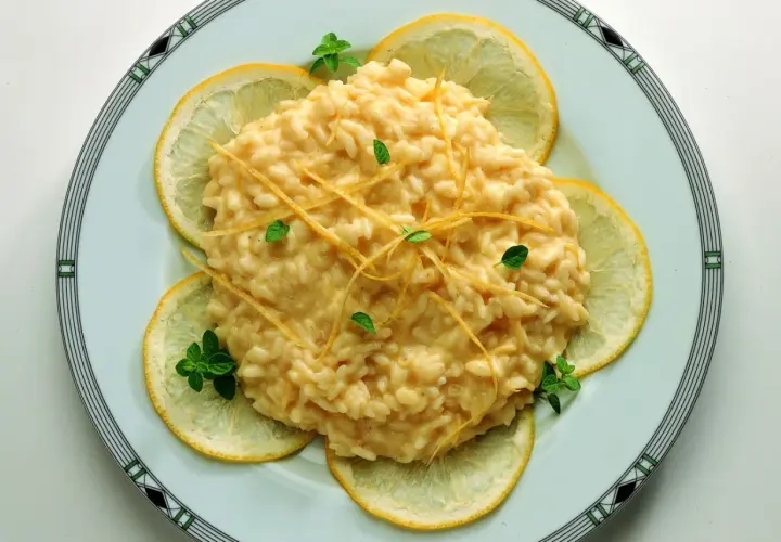Un cremoso risotto al limone, guarnito con scorzette e foglioline di menta, circondato da fette di limone fresco.