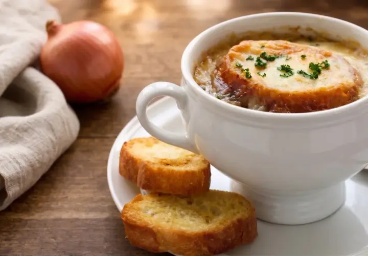 Una tazza di zuppa di cipolle fumante, con crostini dorati e formaggio fuso, accanto a una cipolla cruda e un tovagliolo di lino.