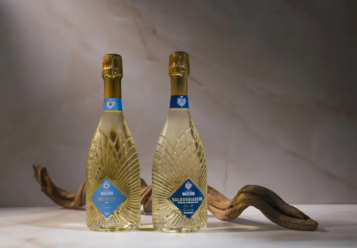 Due bottiglie di Prosecco Maschio, una DOC Treviso e l'altra Valdobbiadene DOCG, con un ramo secco. Scopri come si fa il Prosecco.