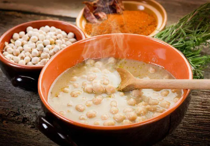Una fumante zuppa di ceci in una ciotola arancione, con ceci secchi, spezie e rosmarino sullo sfondo.