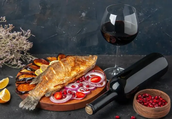 Un delizioso pesce fritto su un piatto con melanzane, cipolle rosse e pomodorini, accompagnato da vino rosso.