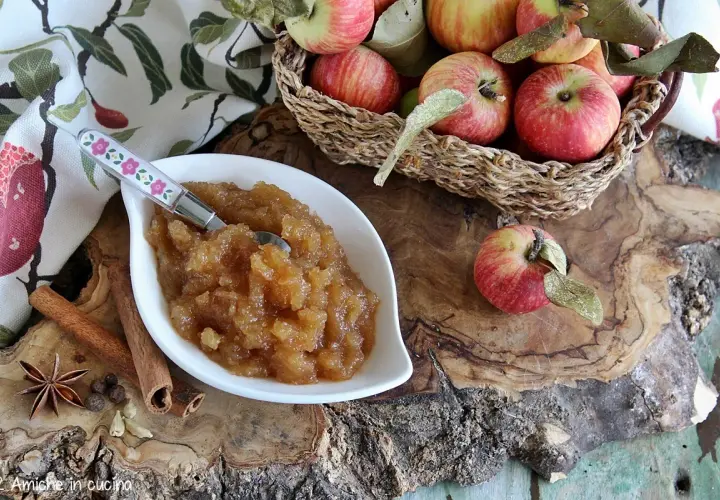 Una ciotola di composta di mele, profumata con cannella e anice stellato, perfetta per la marmellata di natale. Mele rosse in un cesto sullo sfondo.