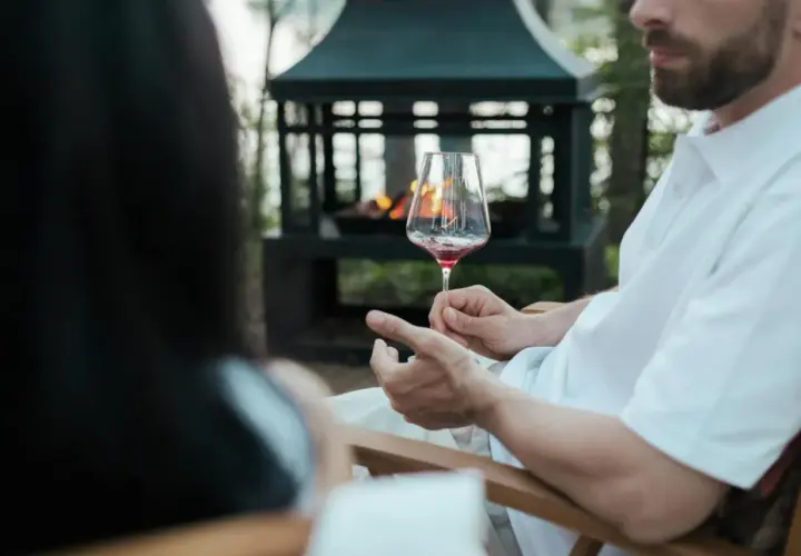 Uomo con barba tiene un calice di vino rosso, mostrando come si tiene il calice di vino per il gambo, vicino a un braciere acceso.