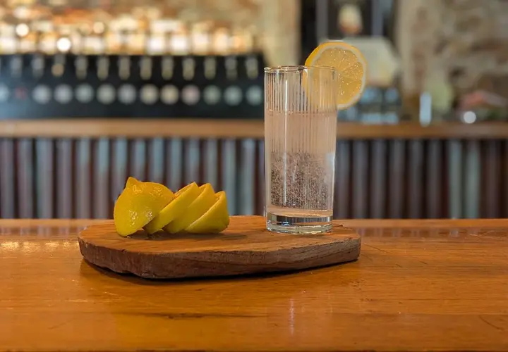 Un bicchiere di acqua frizzante con una fetta di limone, pronto per diventare la base perfetta per un rinfrescante spritz. Accanto, spicchi di limone su un tagliere di legno.