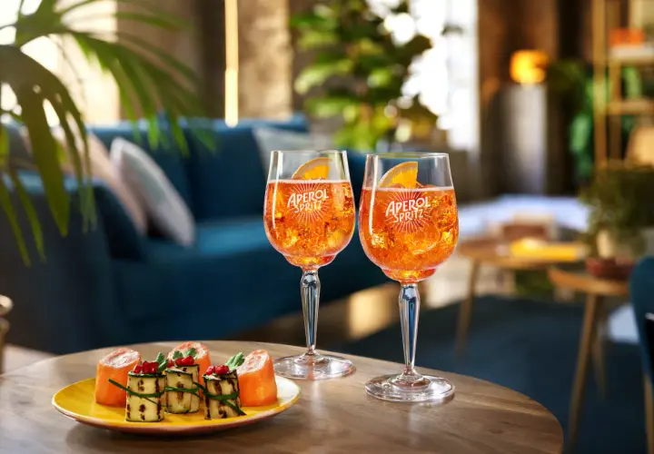Due bicchieri di cocktail con Aperol Spritz, ghiaccio e fetta d'arancia, accompagnati da stuzzichini su un tavolino in legno.