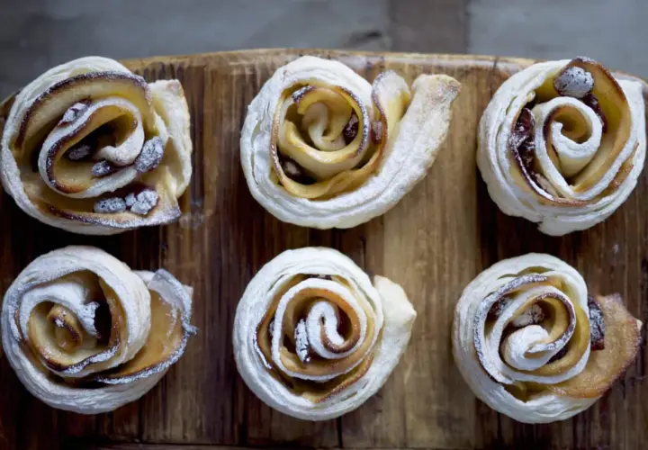 Sei rose di pasta sfoglia con mele e zucchero di canna, cosparse di zucchero a velo, su un tagliere di legno.