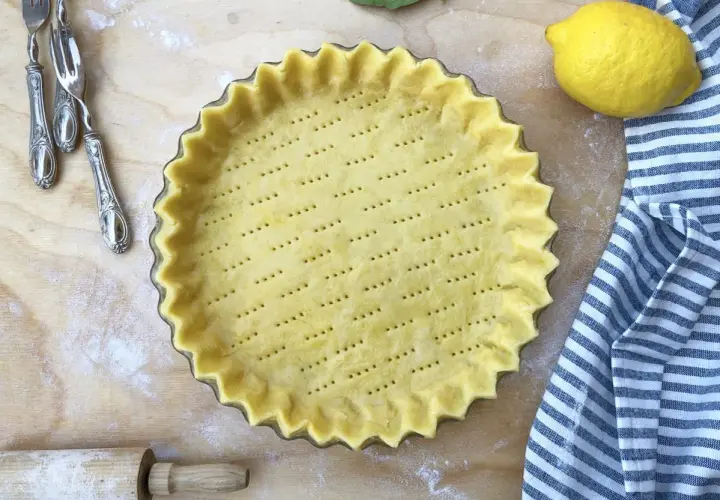 Base di pasta frolla pronta in teglia, forata e con bordi ondulati, accanto a un limone e un canovaccio a righe.