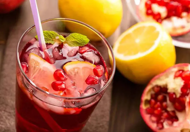 Un rinfrescante spritz al melograno con fette di limone, chicchi rossi e foglie di menta, accompagnato da limoni e melograni freschi.